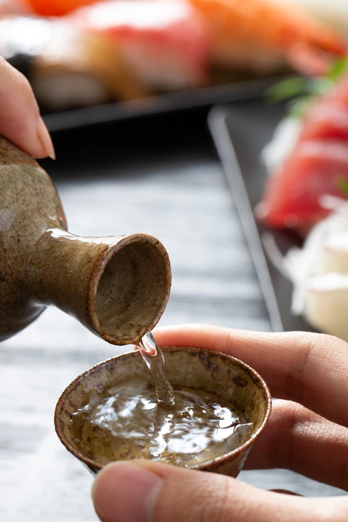 寿司を背景に、徳利から盃へ日本酒を注ぐ手元